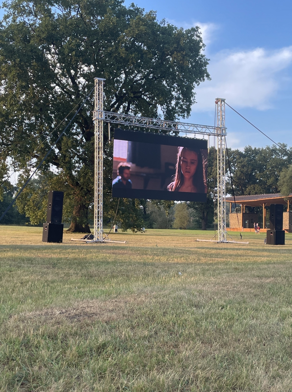 Obraz: Pochmurny dzień. Bytomskie Błonia - łąka w pobliżu Odry, kino w plenerze. Pośrodku ekran projekcyjny, kolumny w foliowych workach, po lewej w namiocie ekipa techniczna, po prawej częściowo widoczni widzowie.